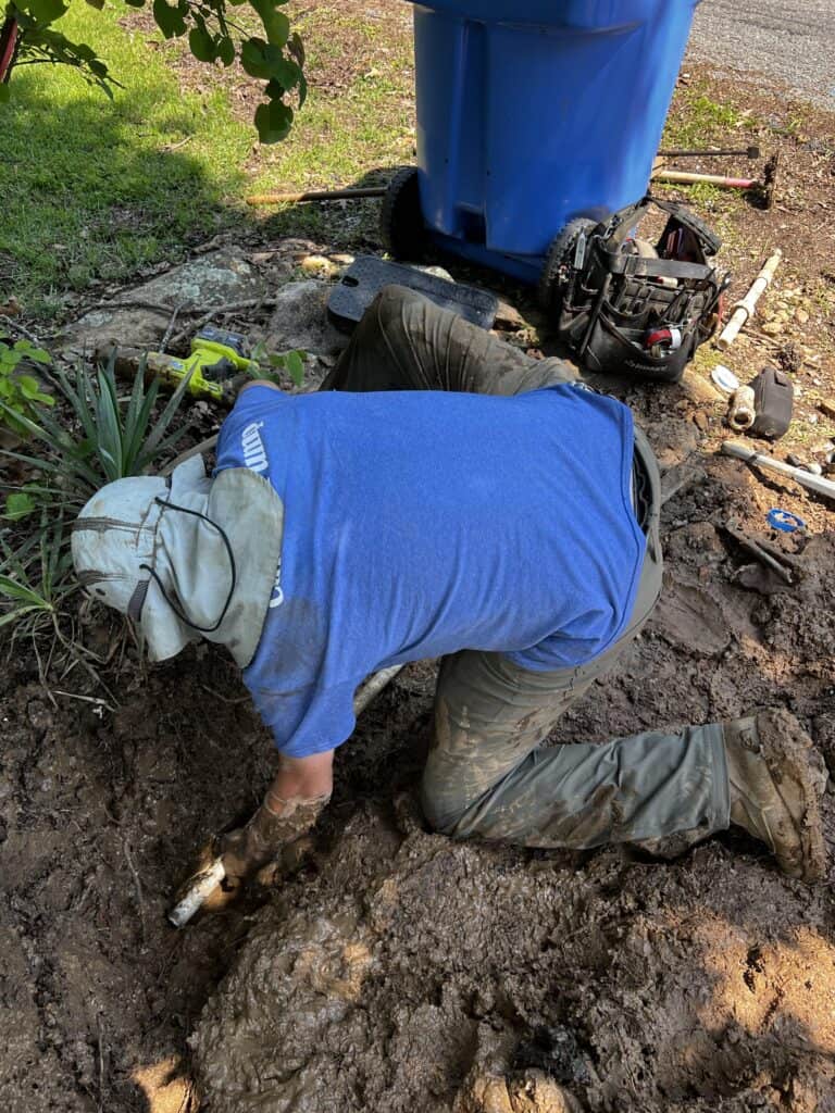 Trench Work - Candor Pump OK Technician in a blue shirt and hat repairing a buried water line in muddy conditions, using tools to access the underground piping.